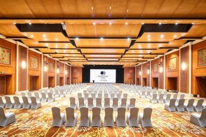 Uma sala de conferências com fileiras de cadeiras, um palco com tela de projeção e painéis de madeira no teto.