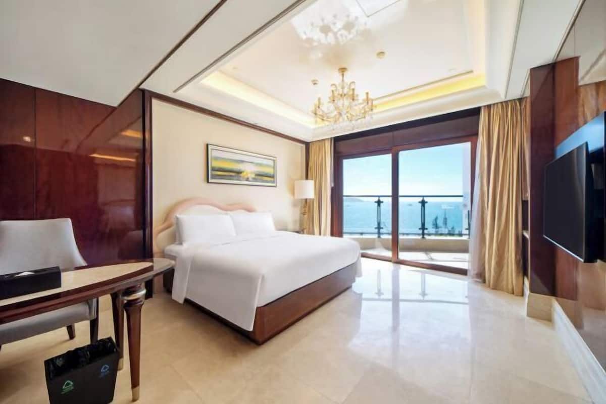 A hotel room with a large bed, a desk, a chair, a TV, a chandelier, a painting, and a balcony with a view.
