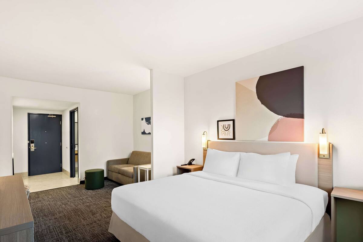 A hotel room with a large bed, a desk, a chair, a small table, and a door.