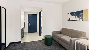 A modern hotel room with a grey sofa, a small round ottoman, a white side table, and a painting on the wall. - Spark by Hilton Williams (Williams)