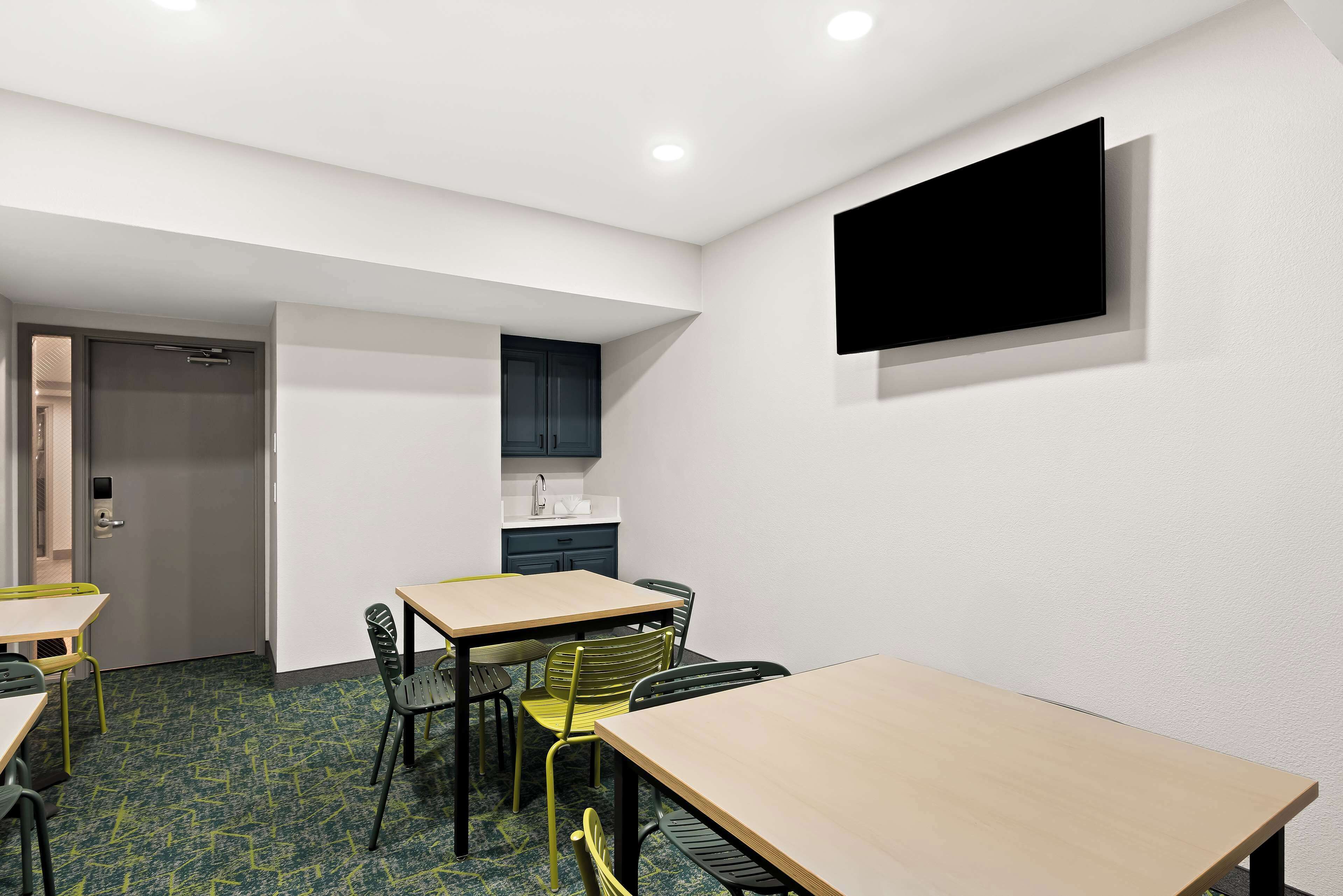 A modern hotel room with a kitchenette, a flat-screen TV mounted on the wall, and a dining area with tables and chairs.