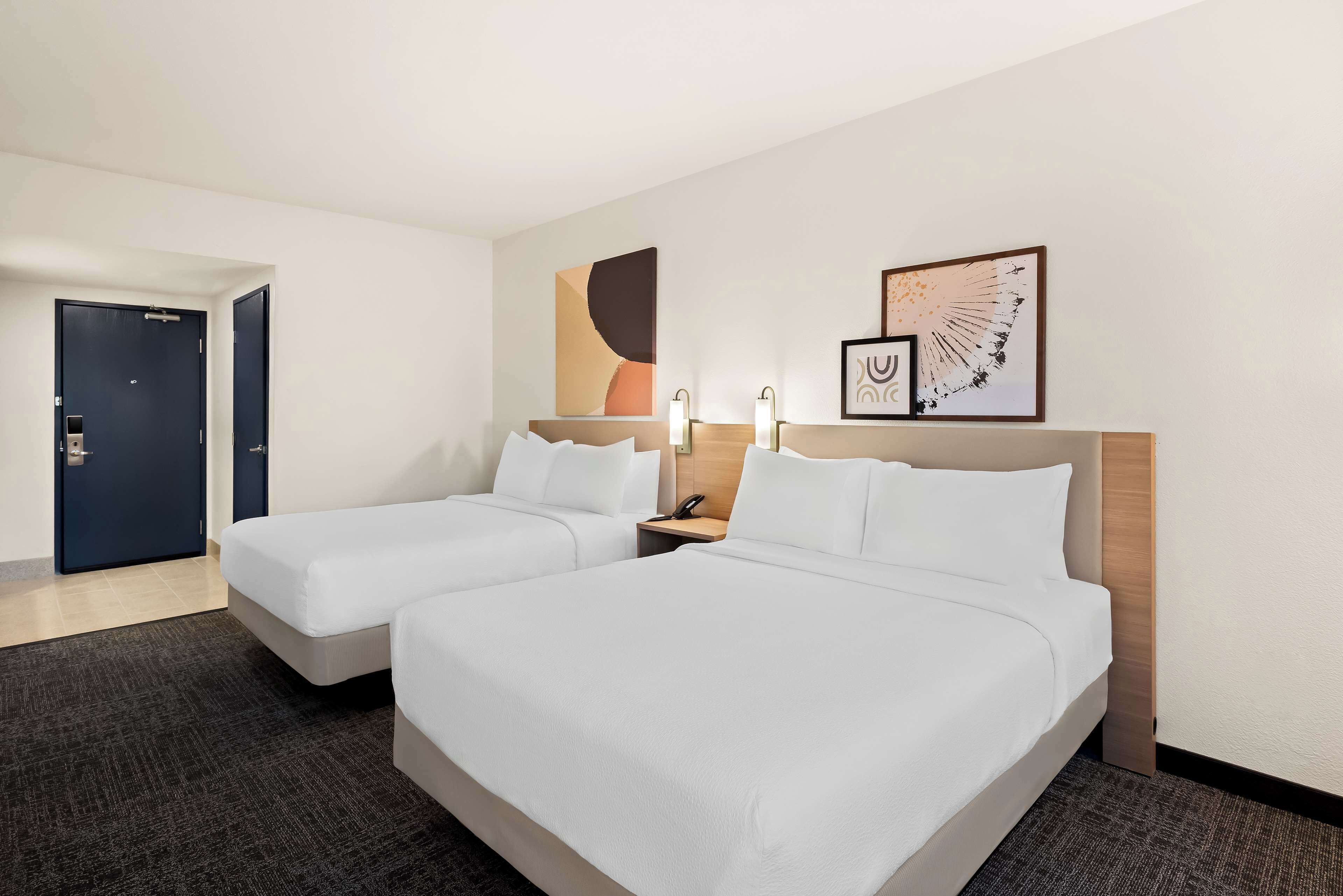 A hotel room with two beds, a dark door, a light-colored wall, and a framed picture.