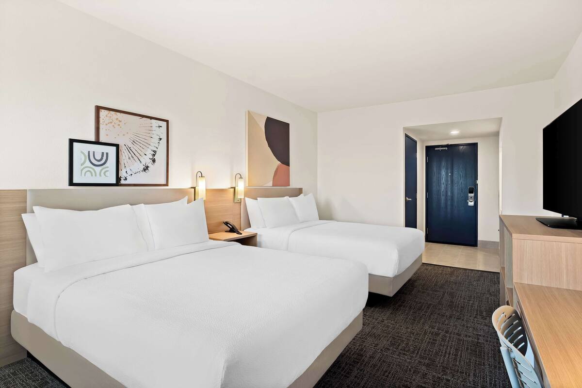 A hotel room with two beds, a desk, a TV, and artwork on the wall.