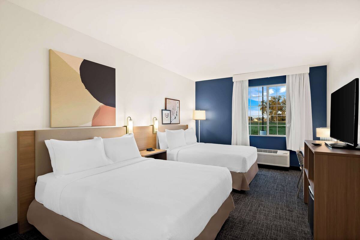 A hotel room with two beds, a desk, a TV, and a window with a view of the outdoors.