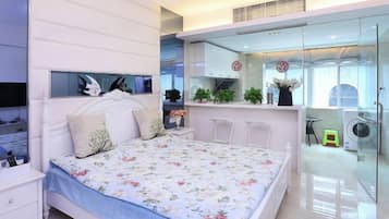 Un dormitorio con cama, mesitas de noche, televisión y un separador de vidrio que divide la habitación de una zona de cocina.