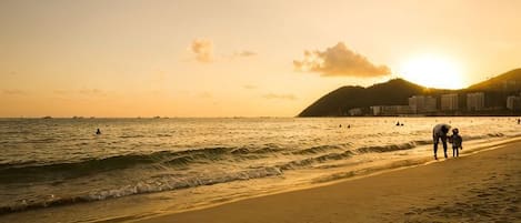 A beach scene with a sunset, waves, and a few people walking along the shore.