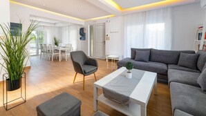 Ein modernes Wohnzimmer mit einem grauen Sofa, einem weißen Couchtisch und einem grauen Sessel. Es gibt einen Essbereich mit einem weißen Tisch und Stühlen sowie ein großes Fenster mit transparenten Vorhängen.