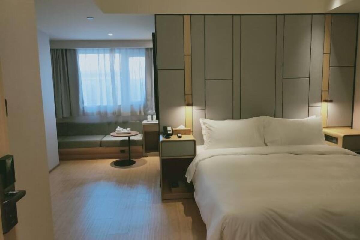 A hotel room with a bed, bedside tables, a nightstand, a small table, a sofa, and a window with curtains.