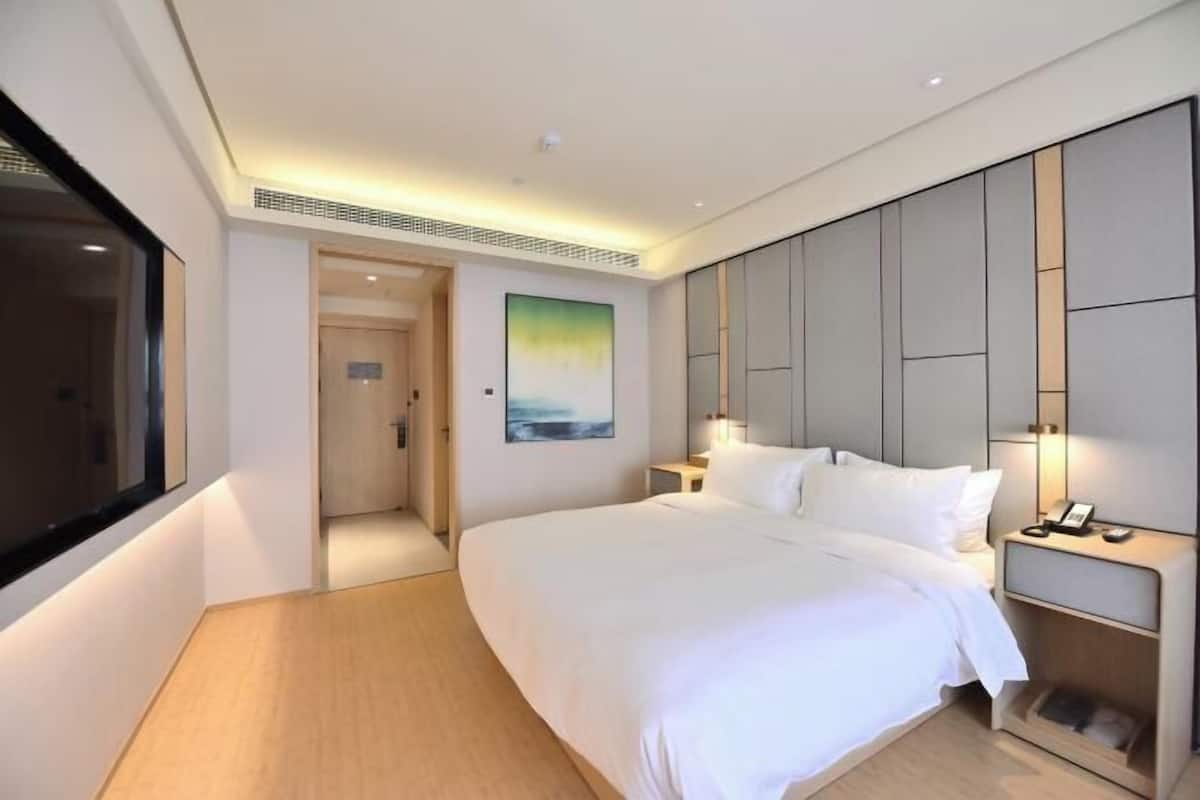 A modern hotel room with a large bed, a bedside table with a lamp, a painting on the wall, and a view of the hallway.