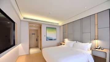 A modern hotel room with a large bed, a bedside table with a lamp, a painting on the wall, and a view of the hallway.