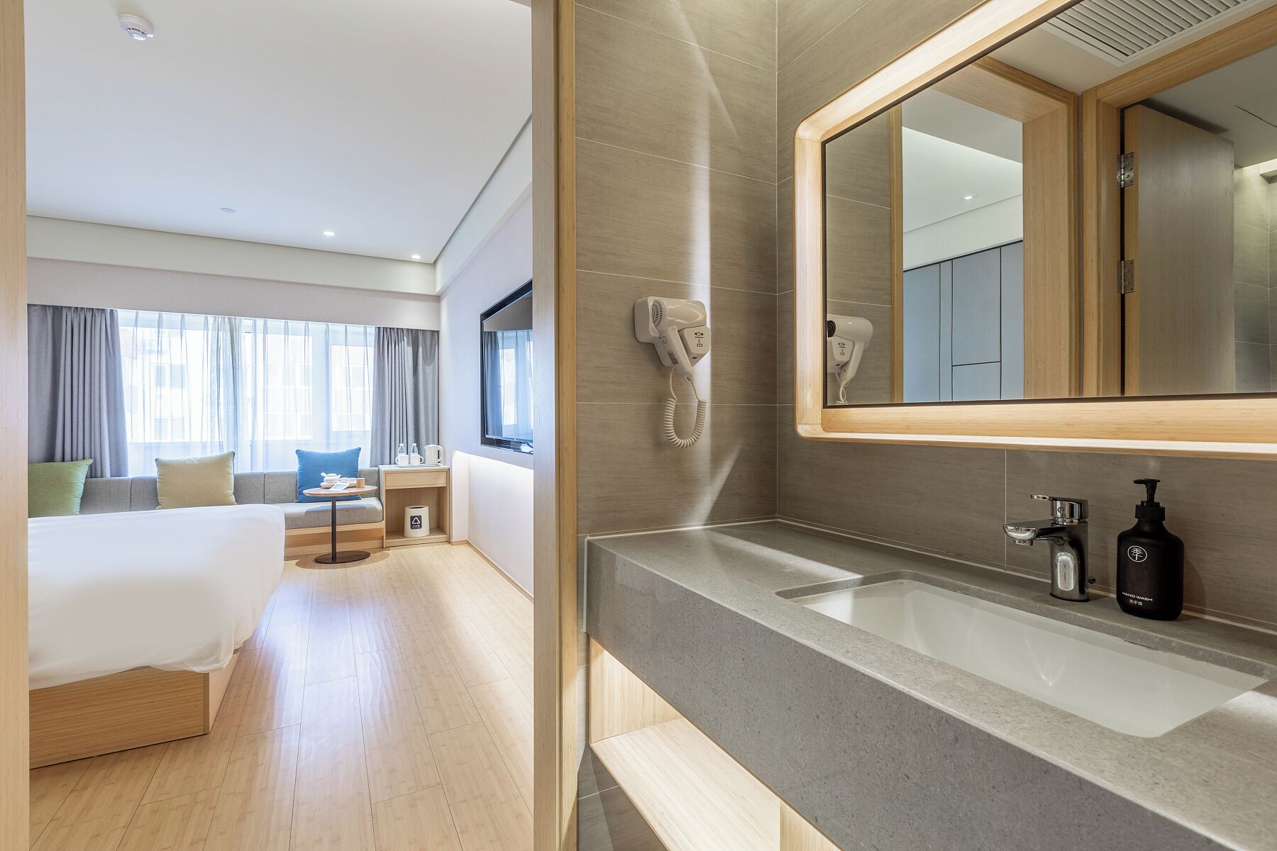 Een moderne hotelkamer met een groot bed, een zithoek met een tafel en stoelen, een badkamer met een grote spiegel en een haardroger aan de muur.