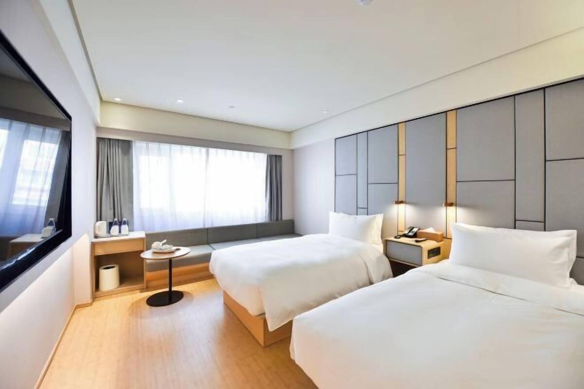 A modern hotel room with two beds, a small seating area, a window with curtains, and a bedside table with a lamp.