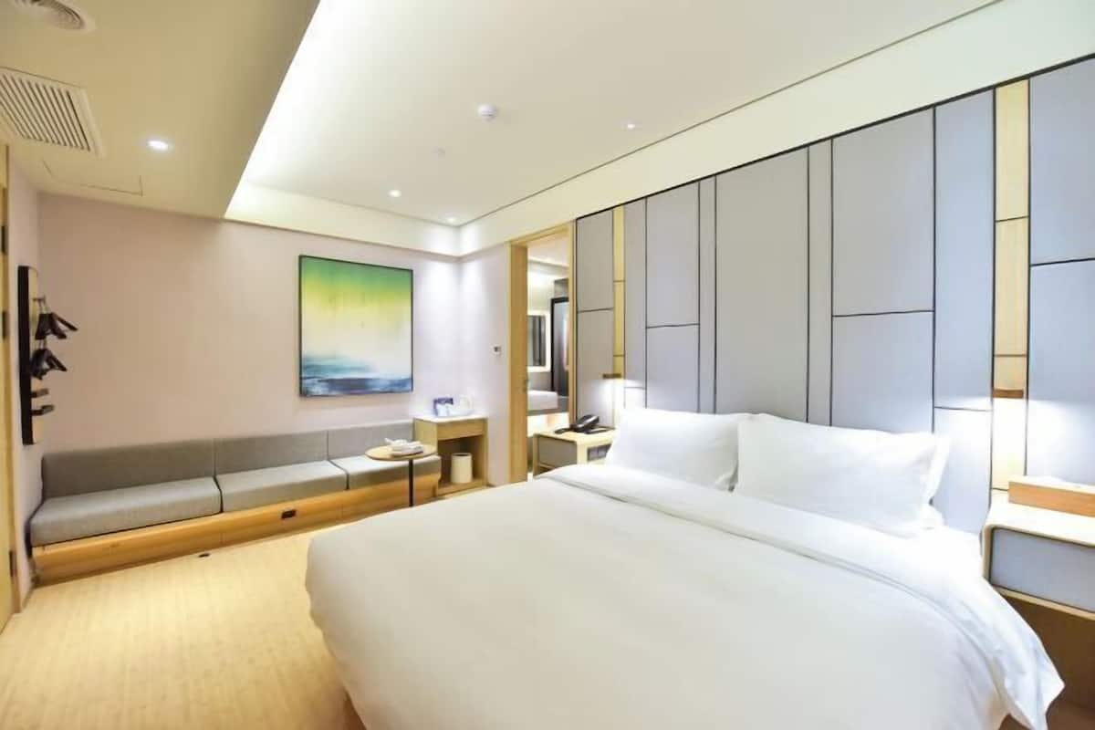 A modern hotel room with a large bed, a seating area, a desk, and a painting on the wall.