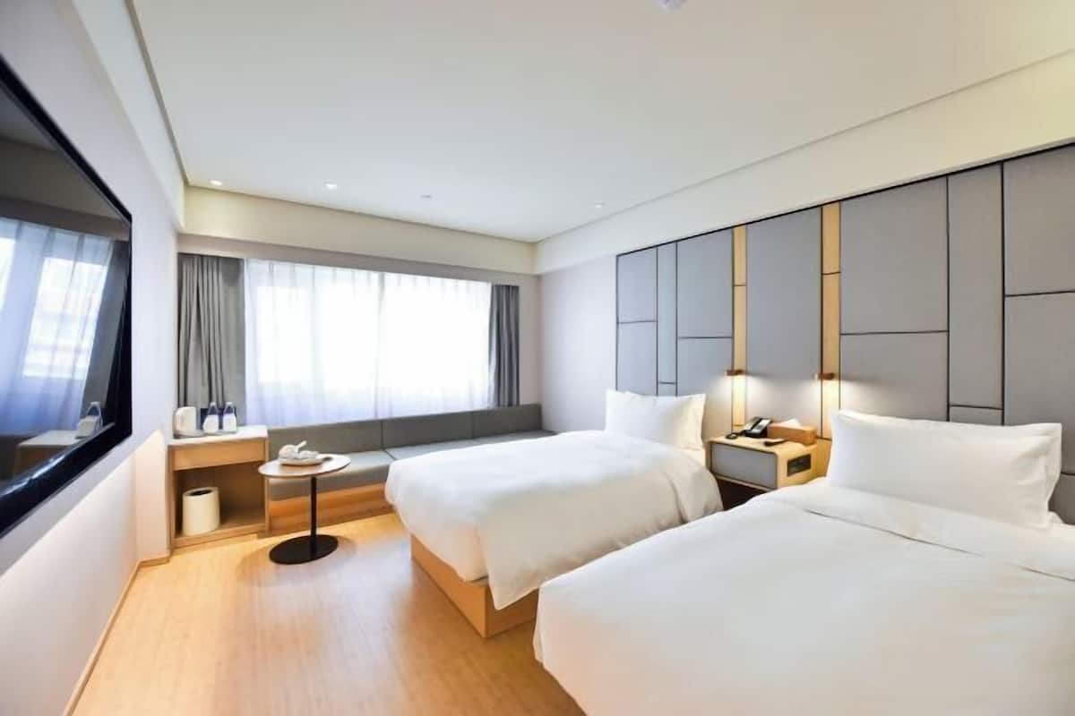 A modern hotel room with two beds, a small seating area, a window with curtains, and a bedside table with a lamp.