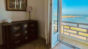 Ein Zimmer mit Holzdielen in Fischgrätmuster, eine dunkle Holzkommode mit verzierten Griffen, ein gerahmtes Bild an der Wand und eine große Schiebeglasdoor zur Terrasse mit Meerblick.