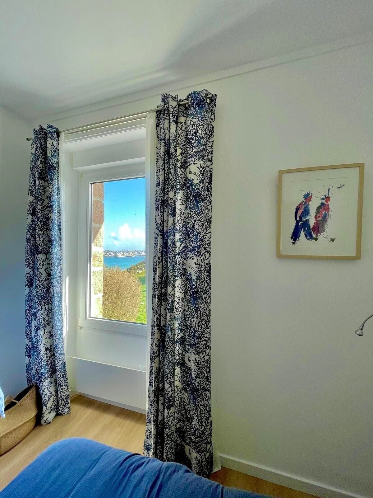 Una habitación con un ventanal grande, cortinas estampadas, una obra de arte enmarcada y una cama.