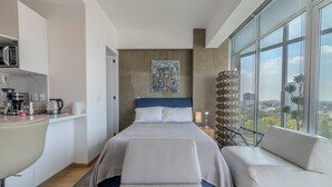 A modern hotel room with a bed, a seating area, a kitchenette, and large windows offering a city view. - Icon Cibeles heart of La Roma (Mexico City)
