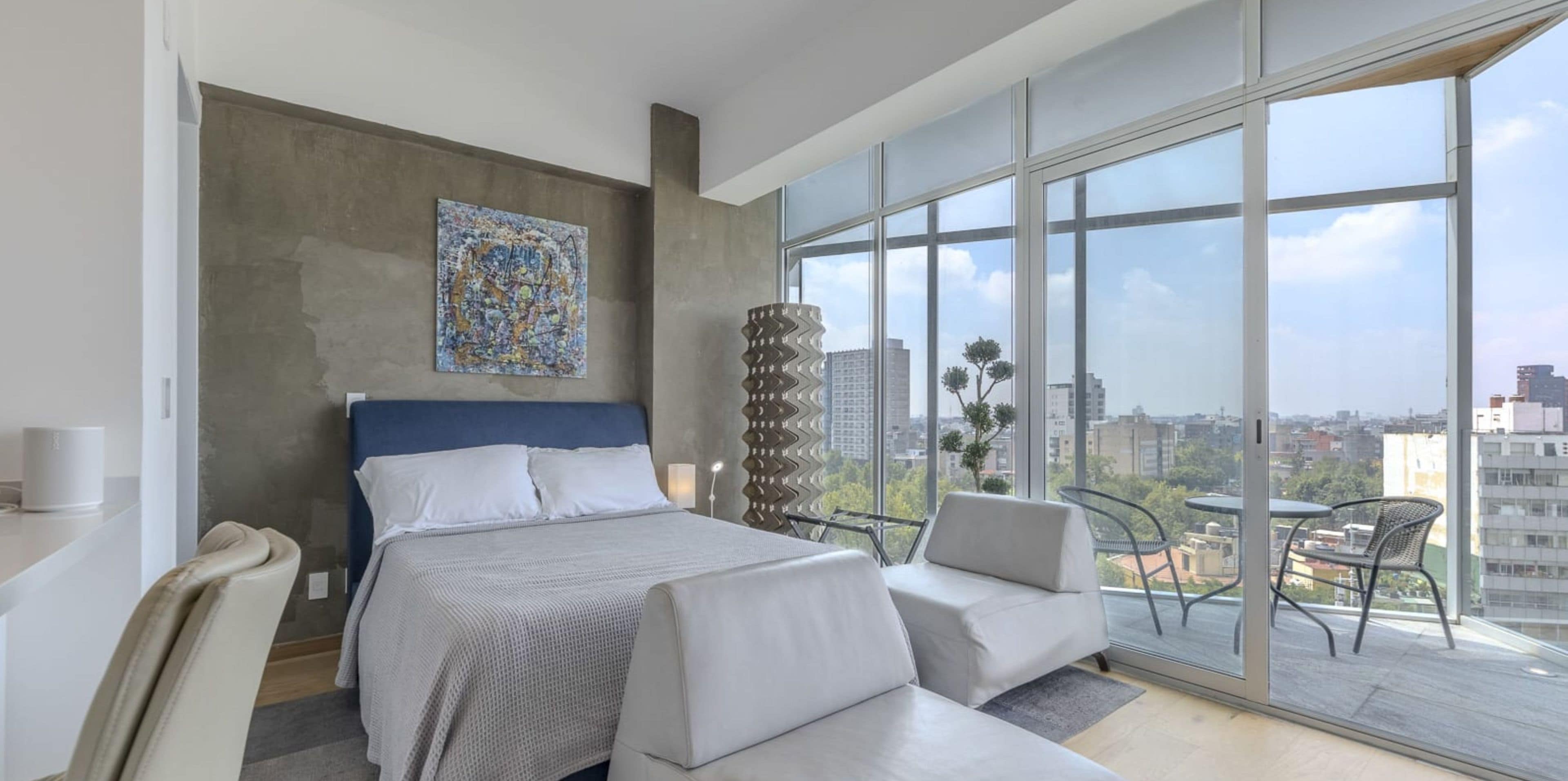 A modern hotel room with a large bed, a sofa, a balcony with a view of the city, and a decorative wall piece.