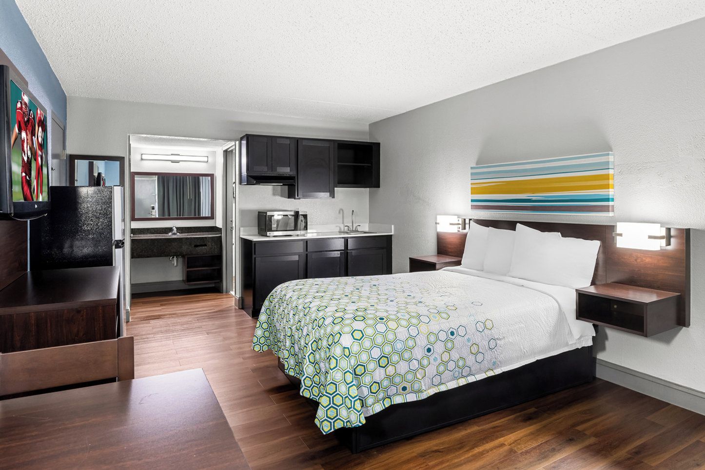 A modern hotel room with a bed, a desk, and a kitchenette.