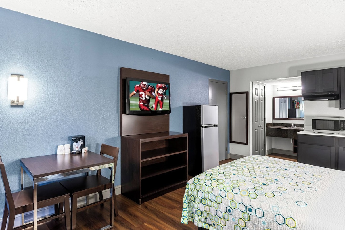 A hotel room with a bed, a TV mounted on the wall, a refrigerator, a microwave, a sink, and a dining table with chairs.