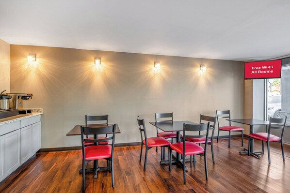 A dining area with a kitchen counter, four tables, and chairs with red cushions. - HomeTowne Studios by Red Roof Richmond - Airport/Sandston (Sandston)