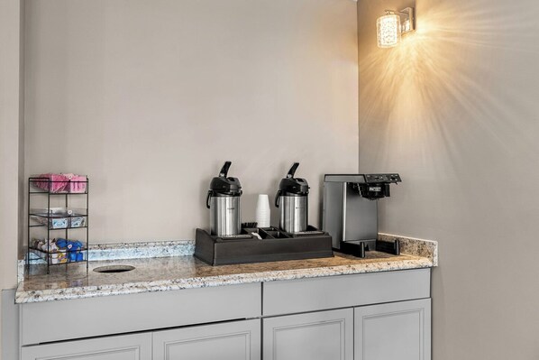 A kitchenette with a coffee maker, two thermos flasks, a coffee dispenser, and a small shelf with cups and a box. - HomeTowne Studios by Red Roof Richmond - Airport/Sandston (Sandston)