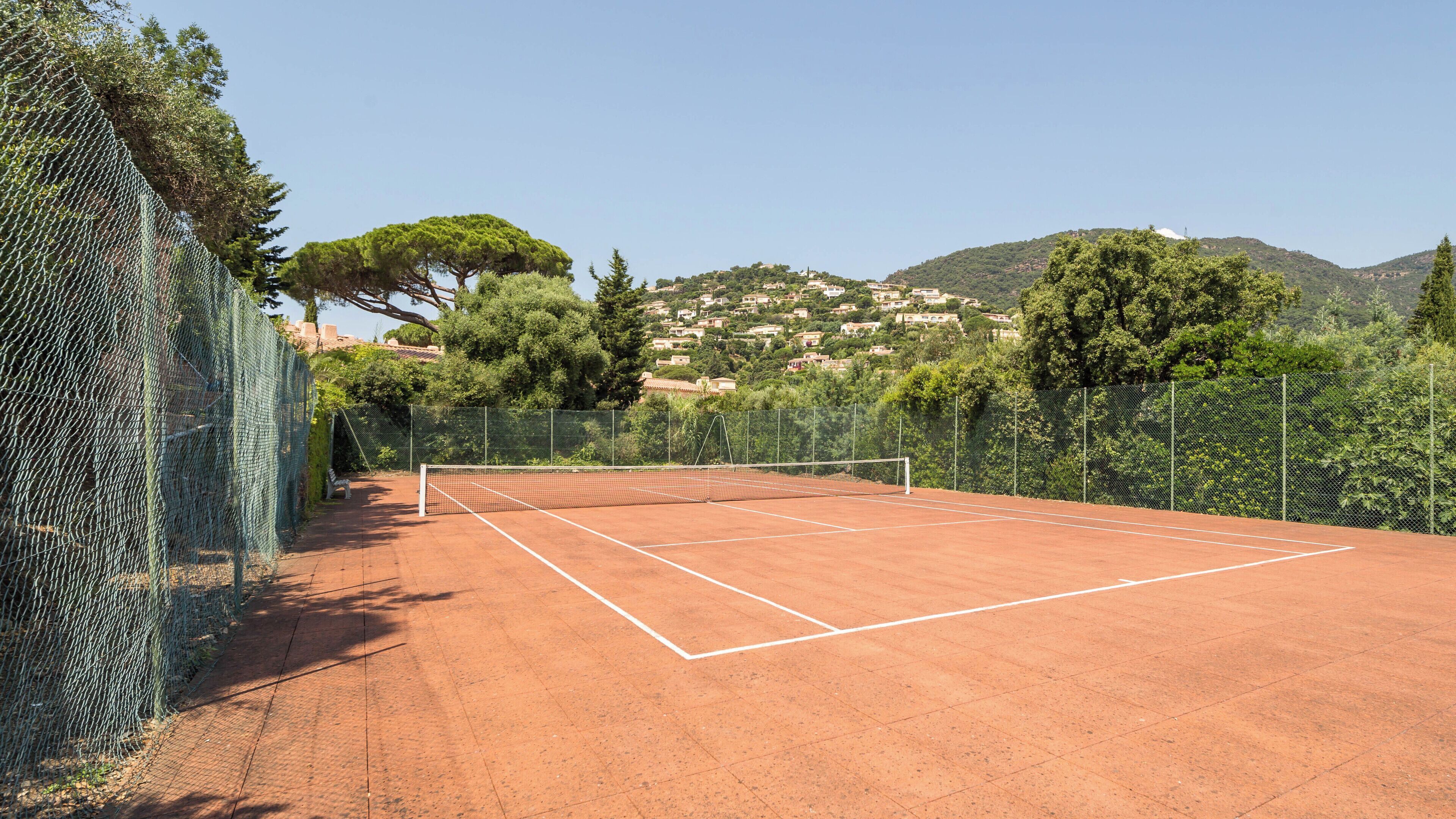 A tennis court with a net, surrounded by a chain-link fence and trees.