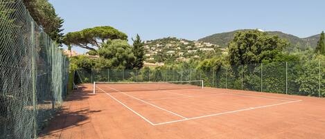 A tennis court with a net, surrounded by a chain-link fence and trees.