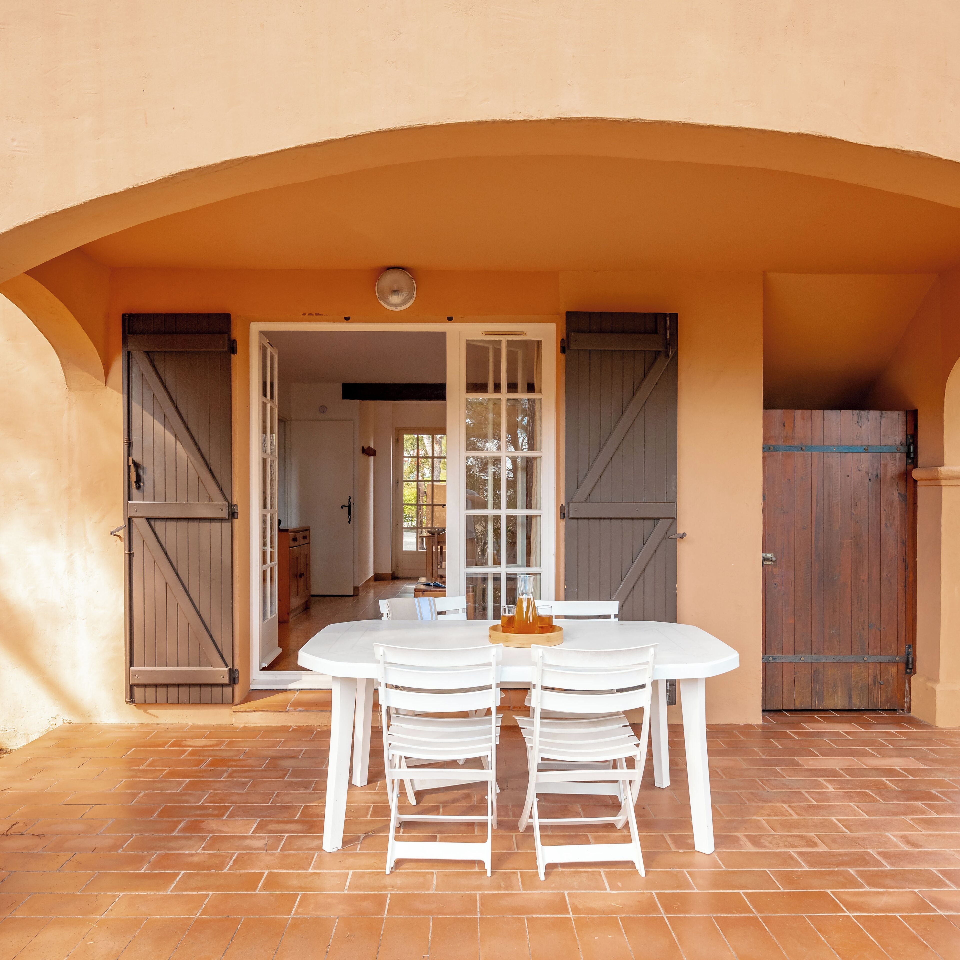 An outdoor patio with a white table and chairs, a wooden door, and a view into the house.