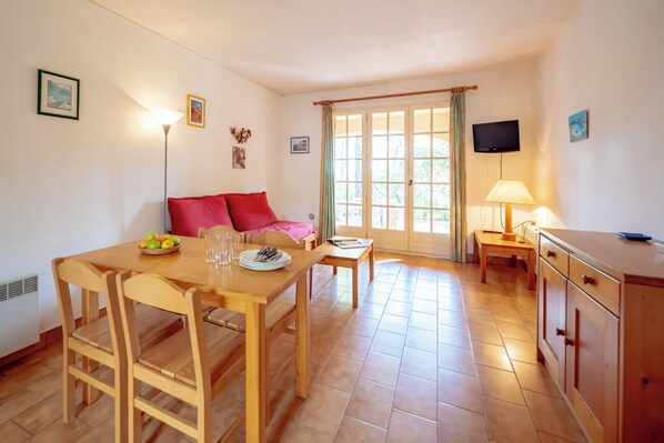 A dining area with a wooden table, chairs, a sofa with red cushions, a television, and a window with curtains. - Résidence Le Domaine de Garlande (Cavalaire-sur-Mer)