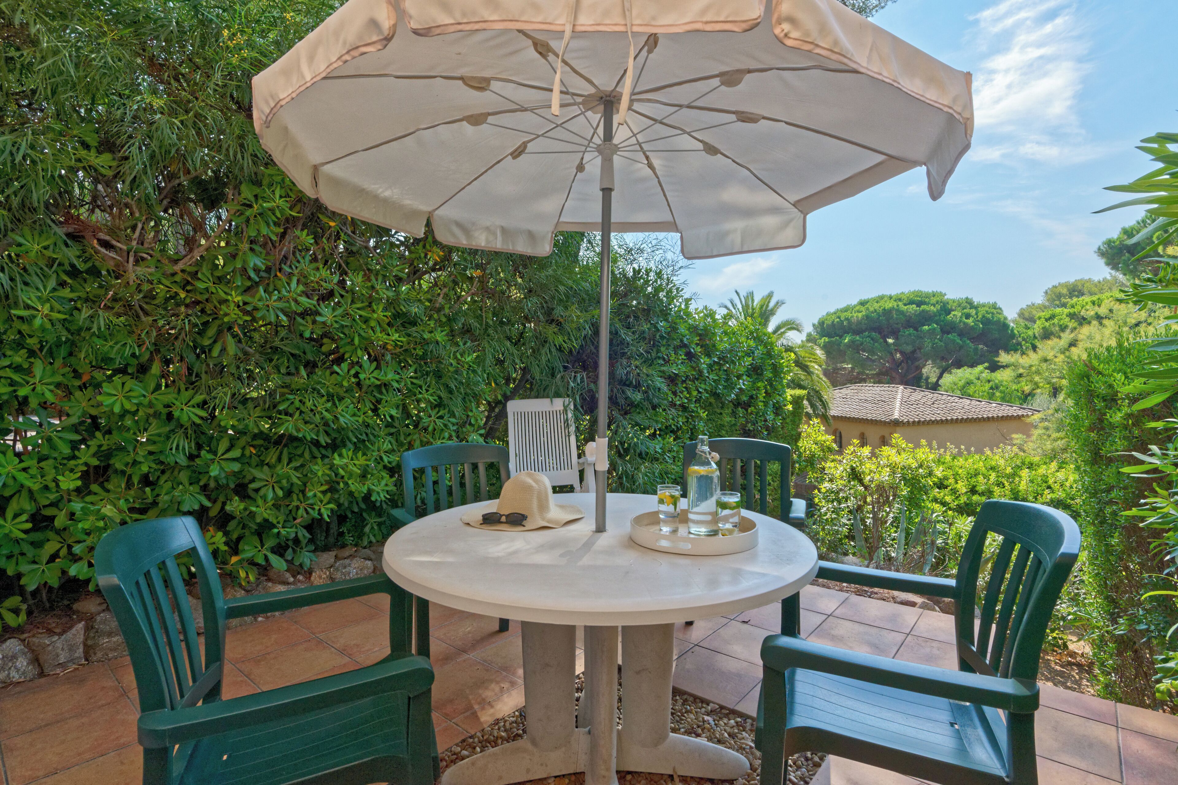 A round table with a beige umbrella, surrounded by green chairs, set on a patio with a view of lush greenery and a house in the background.