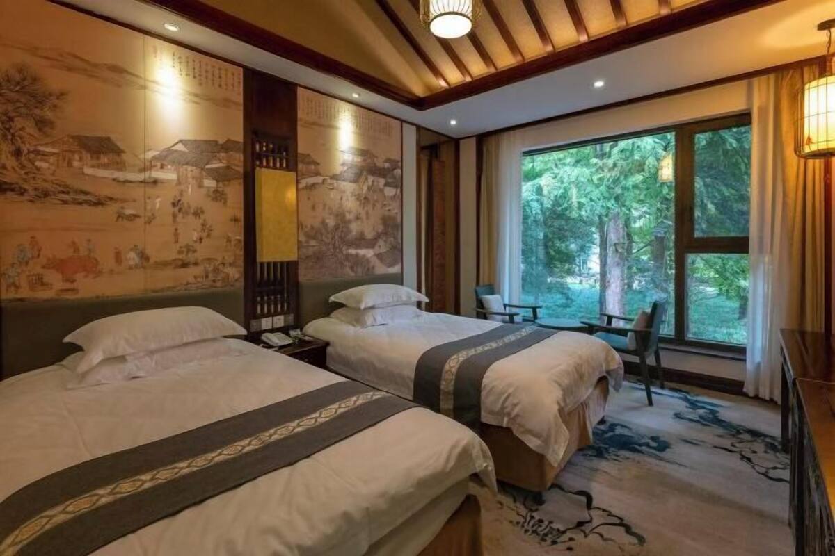 Une chambre d’hôtel avec deux lits, une grande fenêtre donnant sur la verdure et des œuvres d’art traditionnelles accrochées aux murs.