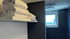 A modern bathroom with a built-in shelf holding folded towels, a window with a view, and a textured wall design. - Dido Diamond Hotel (Dietzenbach)