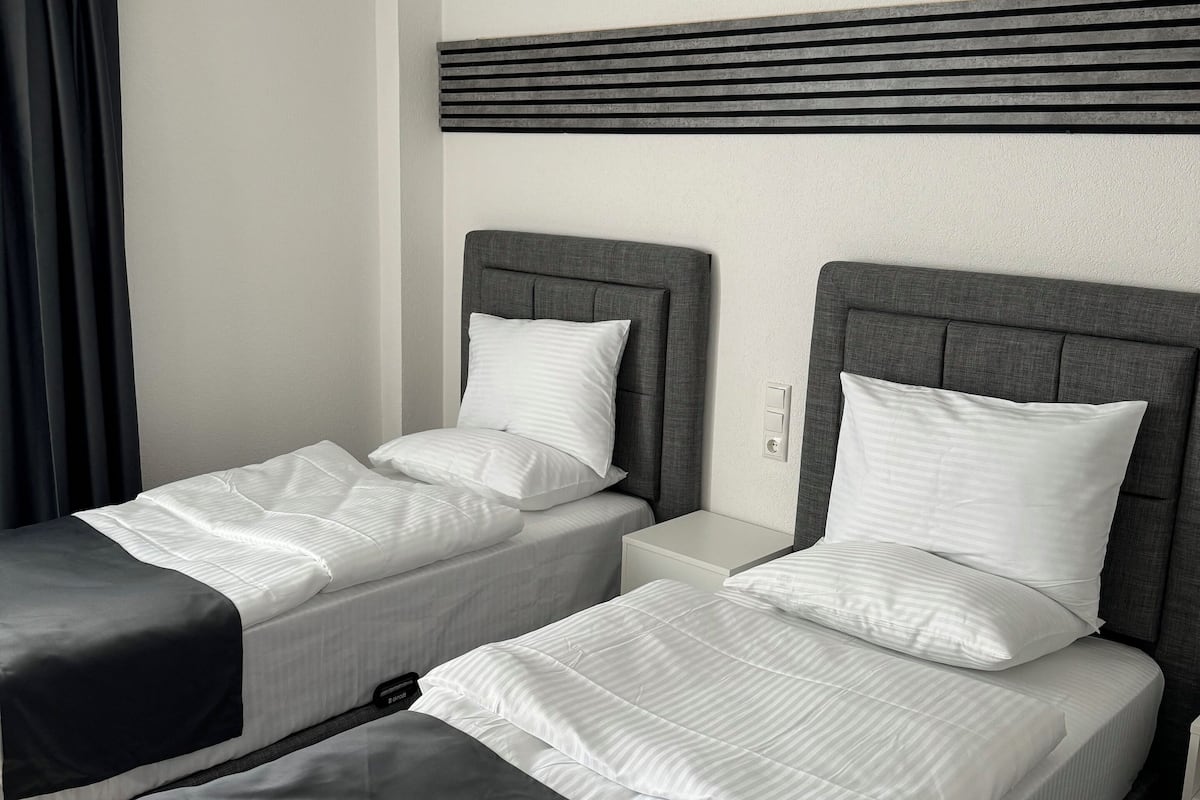 Hotelkamer met twee eenpersoonsbedden, elk met een grijze hoofdbord en witte beddengoed. Er is een moderne plafondlamp en een wandpaneel boven de bedden.