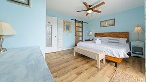 Un dormitorio con cama, piso de madera, ventilador de techo, mesitas de noche y una puerta.