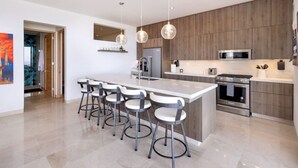 A modern kitchen with a central island, bar stools, stainless steel appliances, and pendant lighting. - Oceanfront 2BR Condo • Panoramic Pacific Views (Los Cerritos)