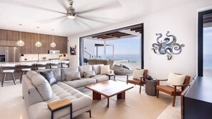 A modern living room with a sectional sofa, coffee table, and a TV stand. There is a kitchen area with wooden cabinets and pendant lights. - Oceanfront 2BR Condo • Panoramic Pacific Views (Los Cerritos)