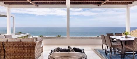 A covered outdoor seating area with a view of the sea, a dining table, and wicker furniture.