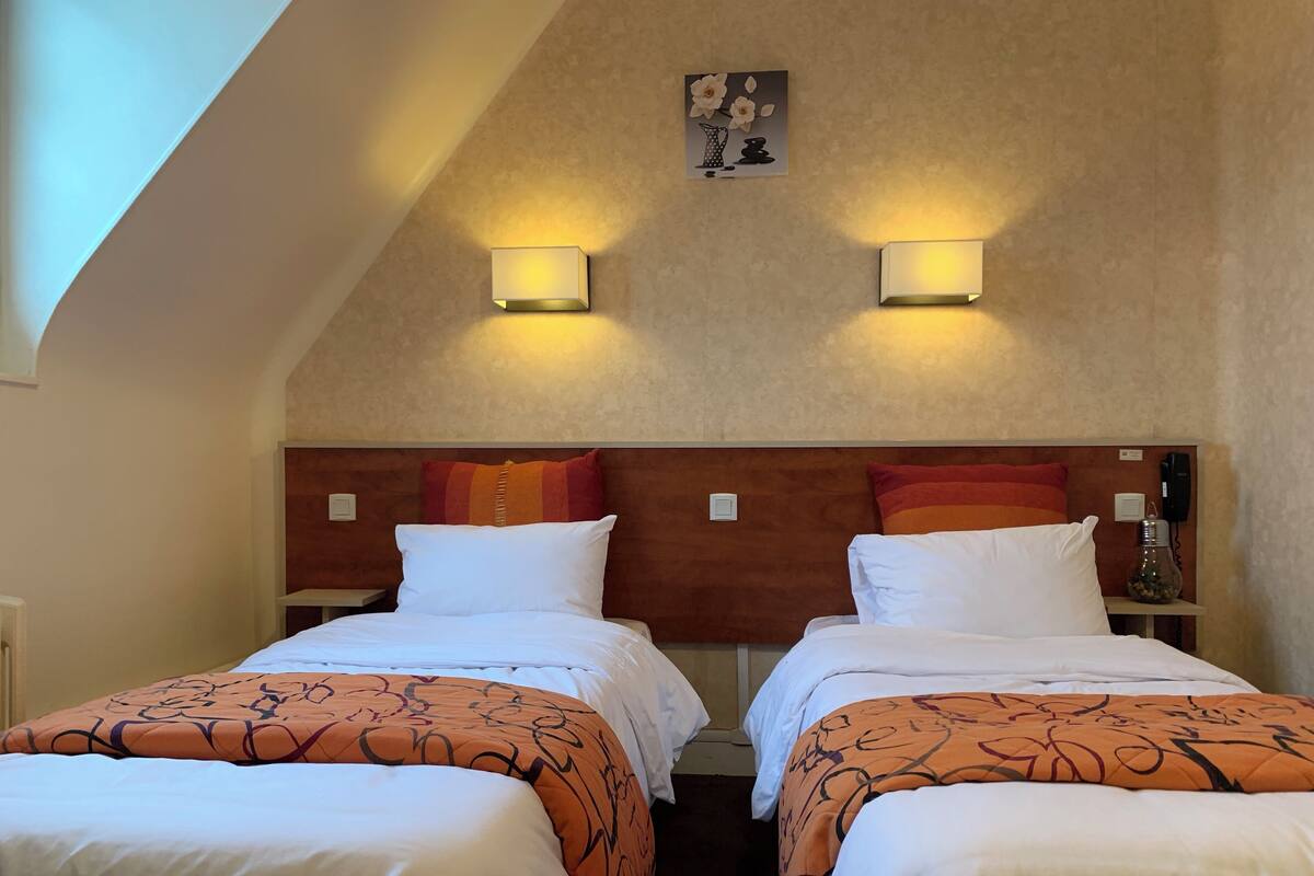 A hotel room with two beds, each with white linens and orange patterned bedspreads. There are two wall-mounted lamps on the ceiling, and a small painting above the beds.