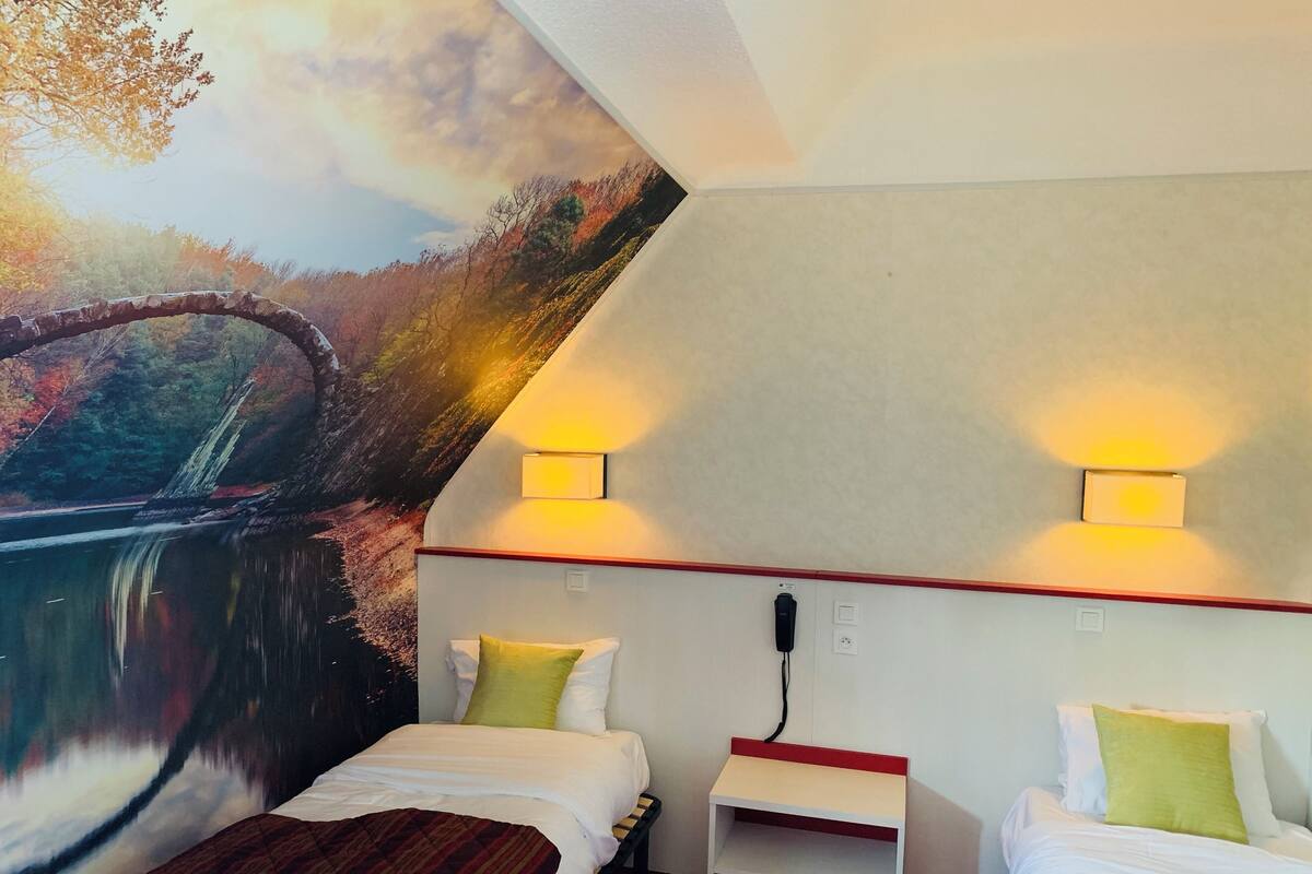 A hotel room with two beds, a small table, and a mural of a bridge over a river.