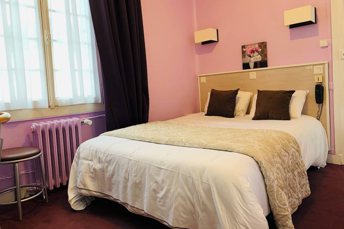 A hotel room with a bed, a chair, a radiator, and a window with curtains.