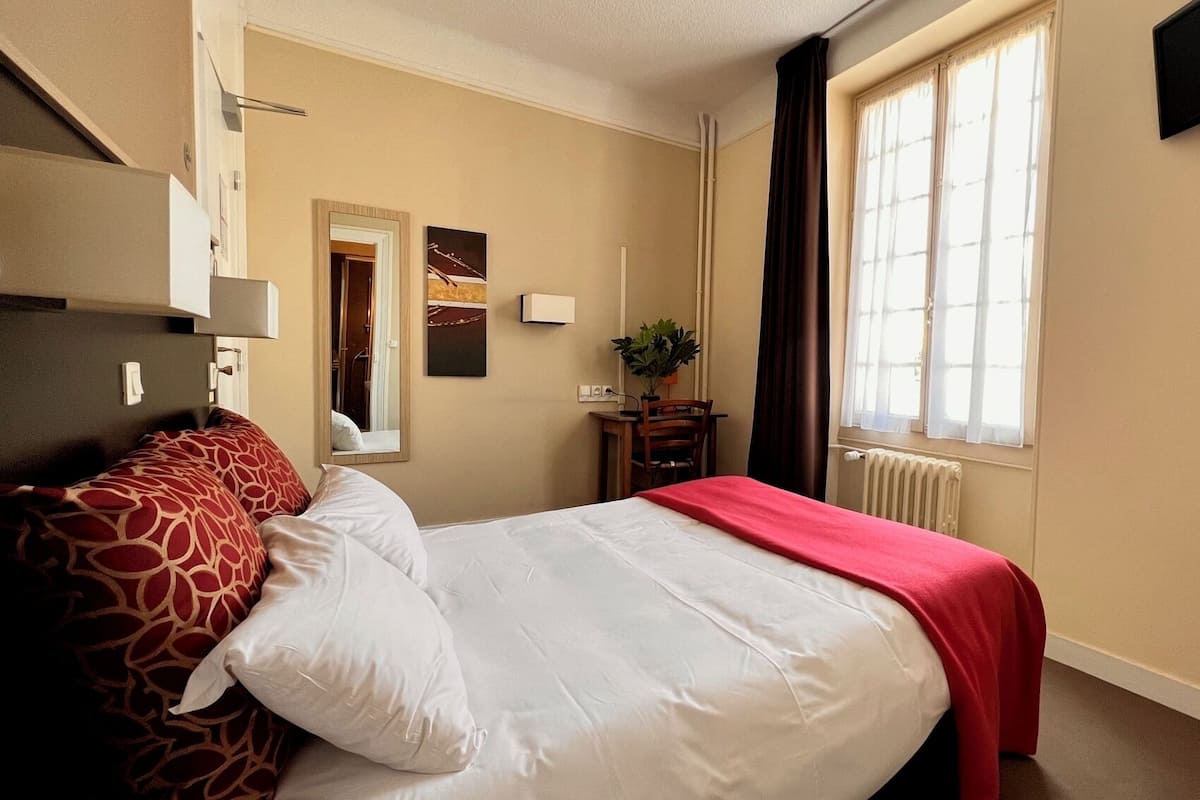 A hotel room with a bed, a desk, a chair, a plant, and a window.