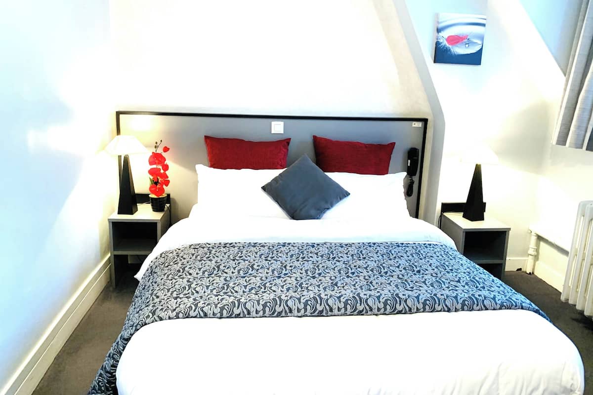 A hotel room with a bed, red and white pillows, a patterned bedspread, a red orchid, a nightstand with a lamp, and a wall-mounted phone.