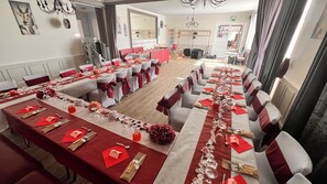 Ein Festsaal mit langen Tischen, gedeckt für eine festliche Veranstaltung, mit roten und weißen Tischdecken, weißen Stühlen mit roten Satinbändern und einer Kristallleuchtre.