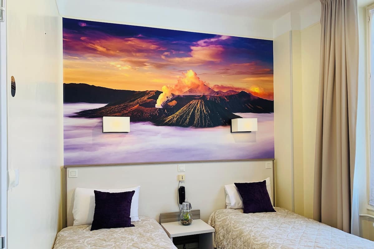 A hotel room with two beds, a large wall mural of a volcano, and a small table with a vase.