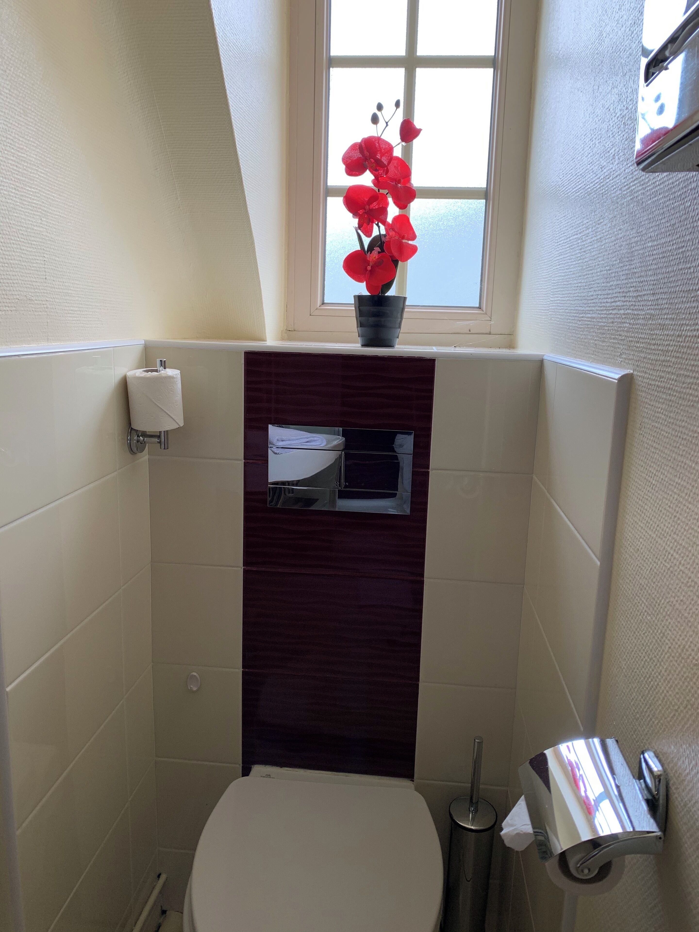 A bathroom with a toilet, a paper towel dispenser, a trash can, and a window with a view of the sky.