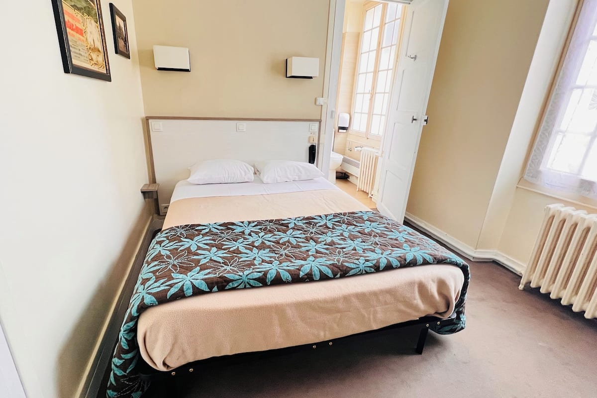 A neatly made bed with a floral bedspread, a white headboard, and a window with a view of the outside.