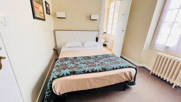 A neatly made bed with a floral bedspread, a white headboard, and a window with a view of the outside.