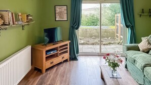A living room with a wooden TV stand, a sofa, a coffee table with flowers, and a large window with curtains. - 2 bedroom accommodation in Wooler (Wooler)