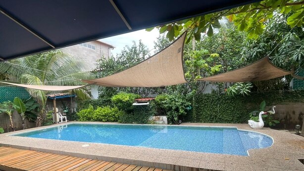 A rectangular swimming pool with a wooden deck surrounding it, shaded by a large canopy.